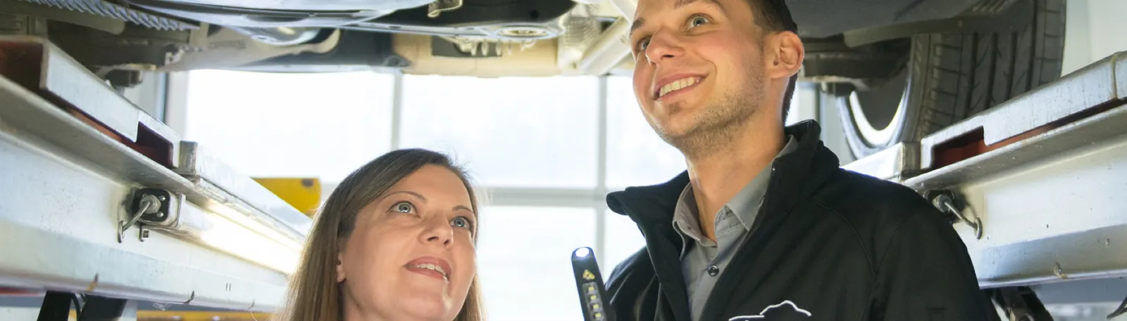 Mitarbeiter der Werkstatt zeigt einer Kundin ihr Auto auf der Hebebühne